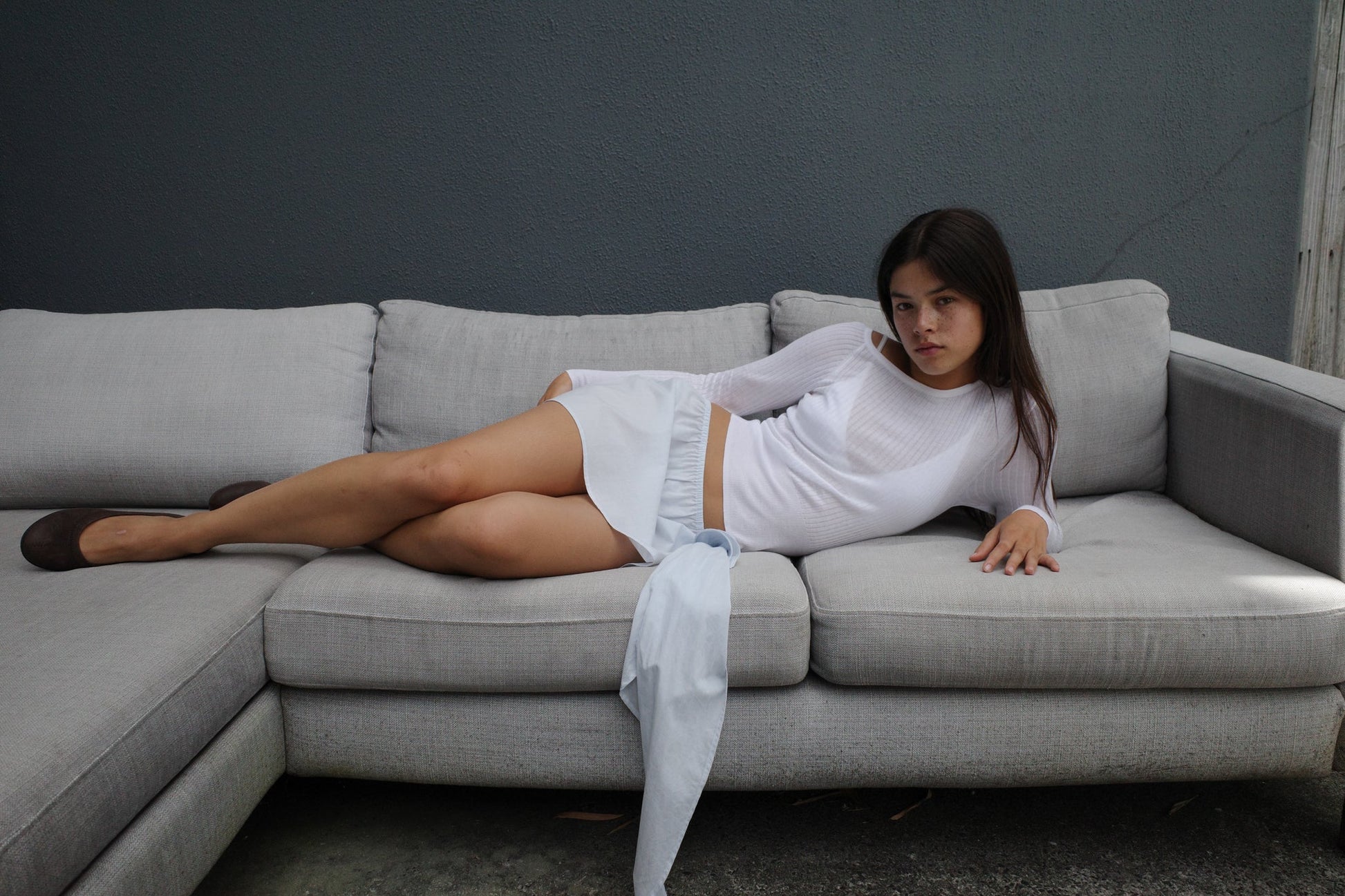 A young woman in a Deiji Studios Shoulder Knit Top - White lounges on a light gray sectional sofa against a dark gray wall, with one arm resting on the back and her legs stretched out on the cushions.