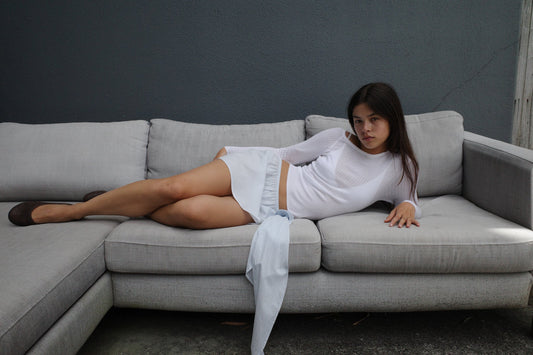A young woman in a white Shoulder Knit Top by Deiji Studios reclines on a light gray sectional sofa against a dark gray wall, gazing toward the camera with a relaxed yet thoughtful expression.