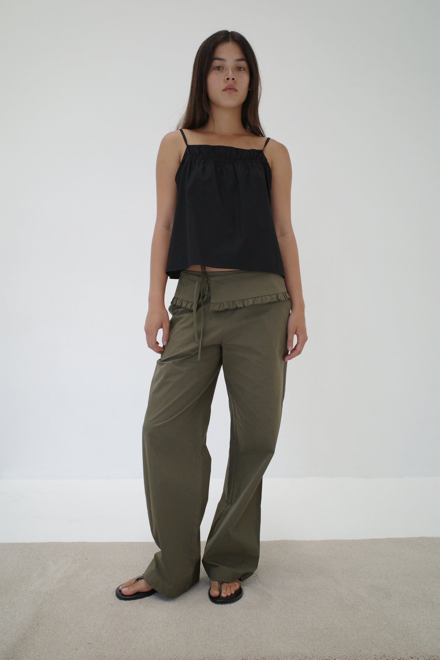 A woman with long dark hair and a neutral expression stands against a white wall wearing a loose black sleeveless top, black sandals, and relaxed fit Flounce Belt Pant - Khaki with ruffle detail and drawstring.