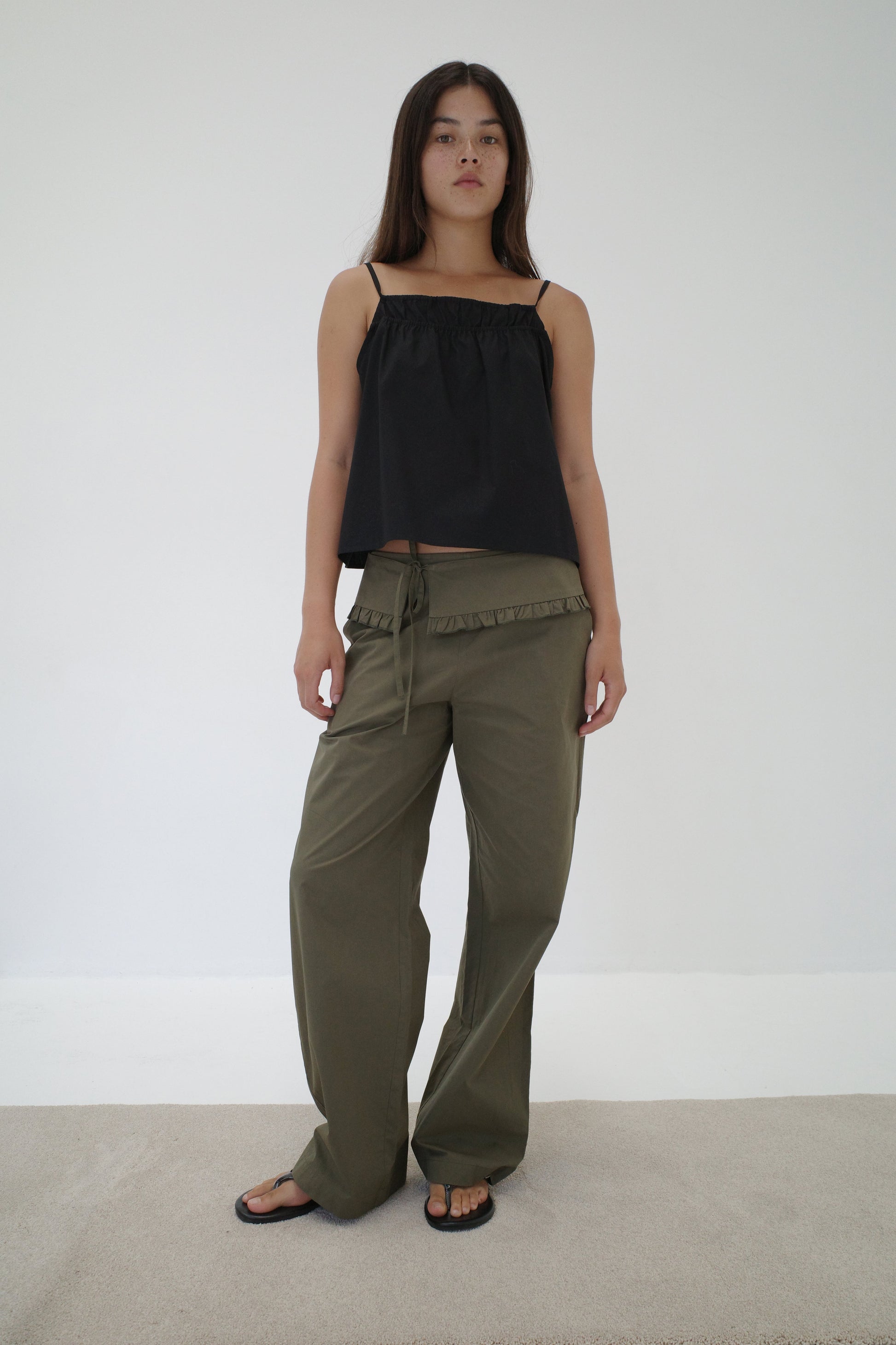 A woman with long dark hair and a neutral expression stands against a white wall wearing a loose black sleeveless top, black sandals, and relaxed fit Flounce Belt Pant - Khaki with ruffle detail and drawstring.