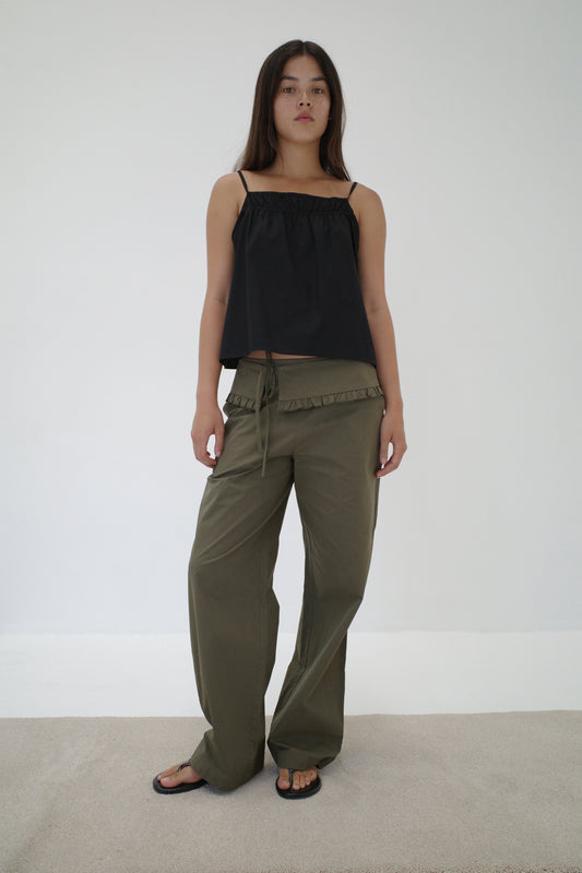 A woman with long dark hair and a neutral expression stands against a white wall wearing a loose black sleeveless top, black sandals, and relaxed fit Flounce Belt Pant - Khaki with ruffle detail and drawstring.