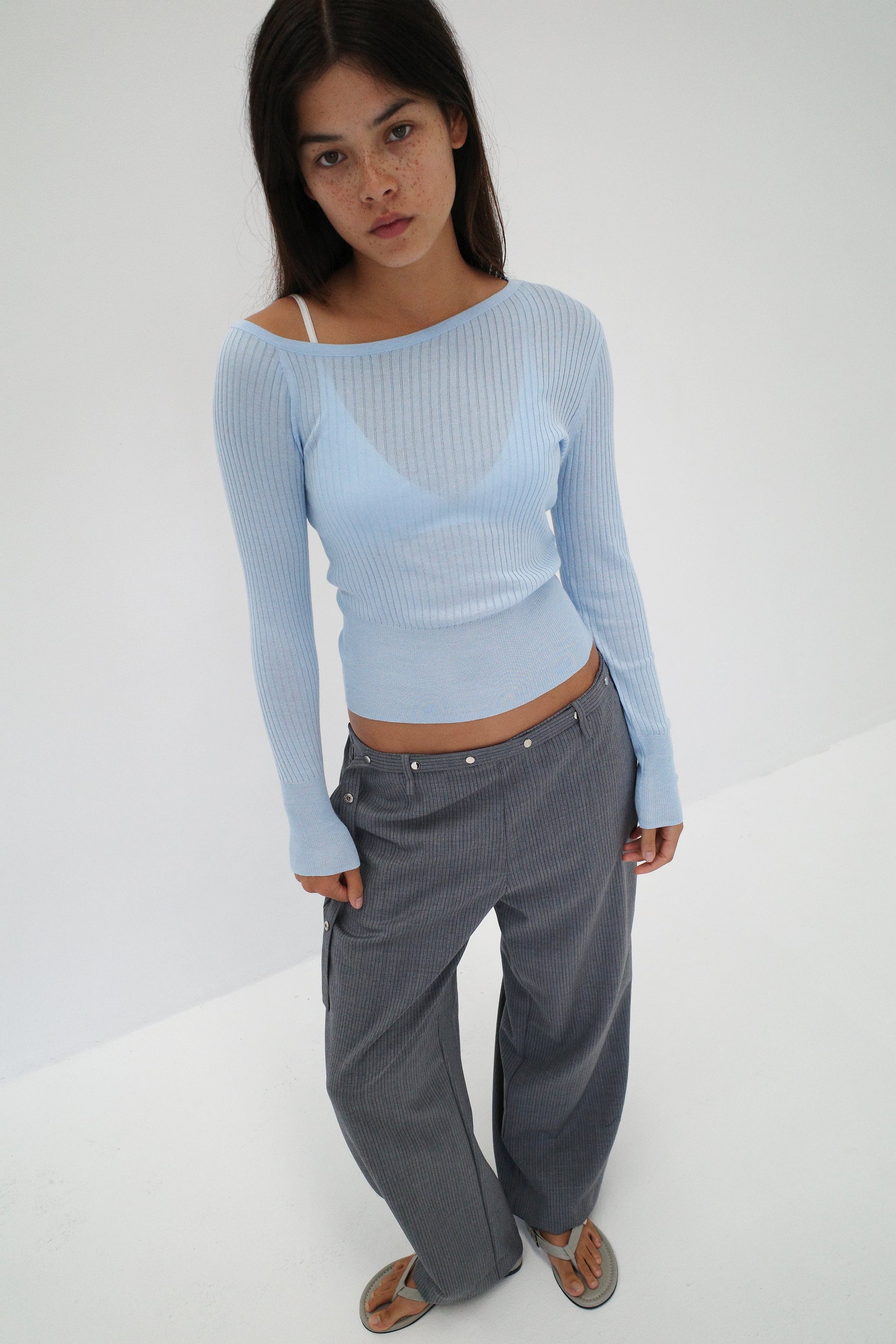 A young woman with long dark hair wears the Shoulder Knit Top - Blue by Deiji Studios, loose gray pinstriped pants, and sandals. She stands against a white background, looking up at the camera with a neutral expression.