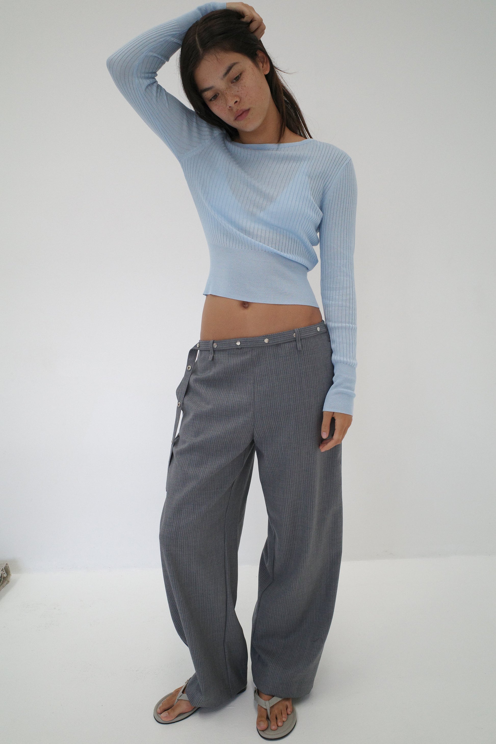 A woman stands against a white background wearing the Shoulder Knit Top - Blue from Deiji Studios and loose gray belted pants. Barefoot, she raises one arm to touch her head and gazes downward.