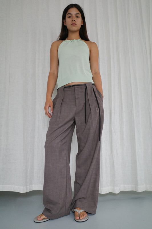 A woman stands before white curtains in a light green halter top, Deiji Studios’ relaxed wide leg Belt Loop Pant in Grey Pink Stripe, a black belt, and gray sandals. She looks confidently at the camera with one hand in her pocket.