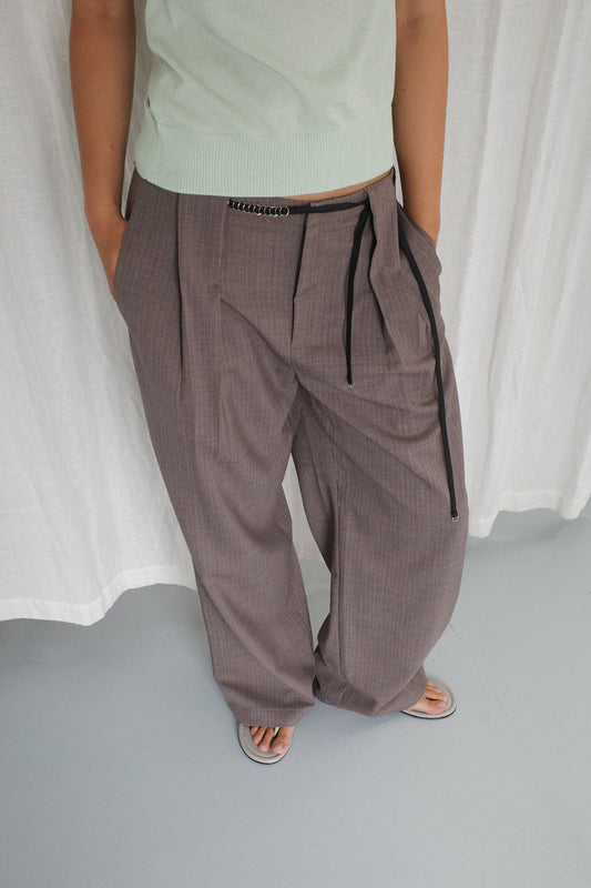 A person wears Deiji Studios' Belt Loop Pant - Grey Pink Stripe with a pale green top and sandals, standing hands-in-pockets against a light background.