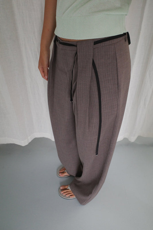 A person wearing Belt Loop Pant - Grey Pink Stripe by Deiji Studios with a black drawstring, a light green top, and beige sandals stands on a light grey floor in front of a white curtain.
