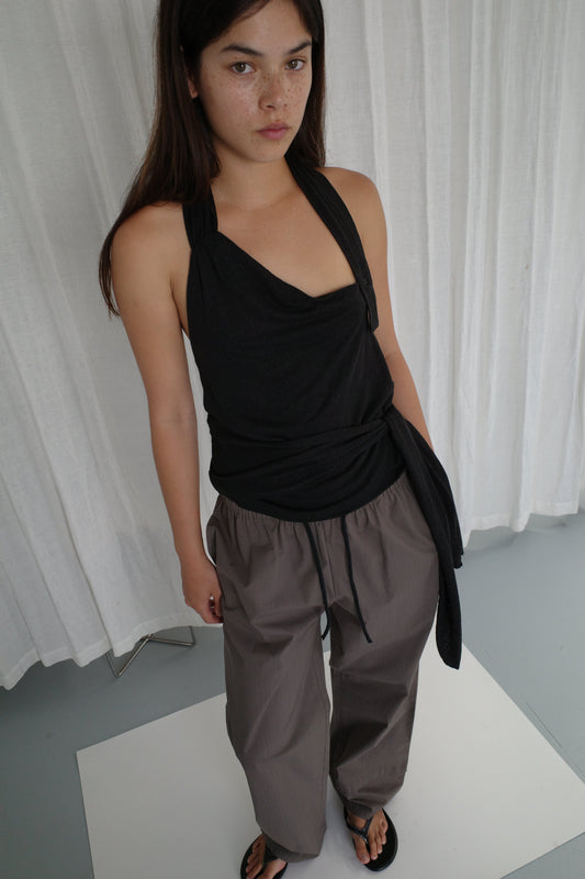 A woman with long dark hair stands indoors in front of white curtains, wearing a sleeveless black halter top, Layered Pant - Khaki Check, and black sandals. She looks slightly up at the camera with a neutral expression.
