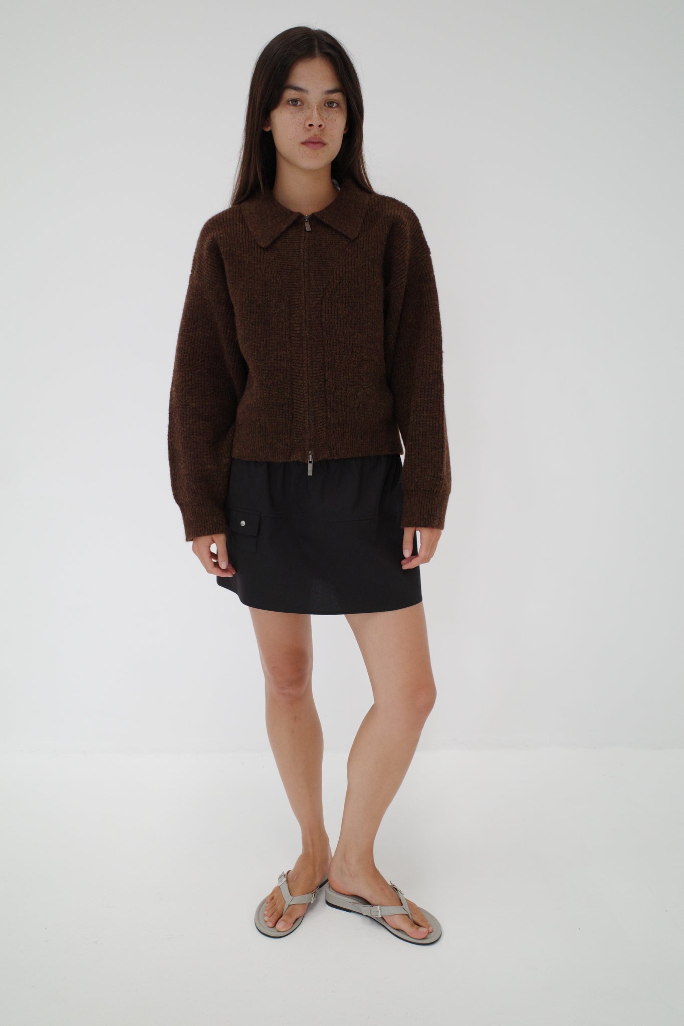 A woman with straight long dark hair and a neutral expression stands against a plain white background wearing Deiji Studios' Double Layer Cardigan in brown, paired with a black skirt and gray flip-flops.