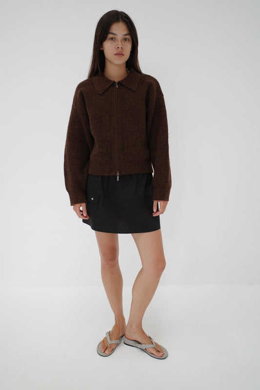 A woman with straight long dark hair and a neutral expression stands against a plain white background wearing Deiji Studios' Double Layer Cardigan in brown, paired with a black skirt and gray flip-flops.