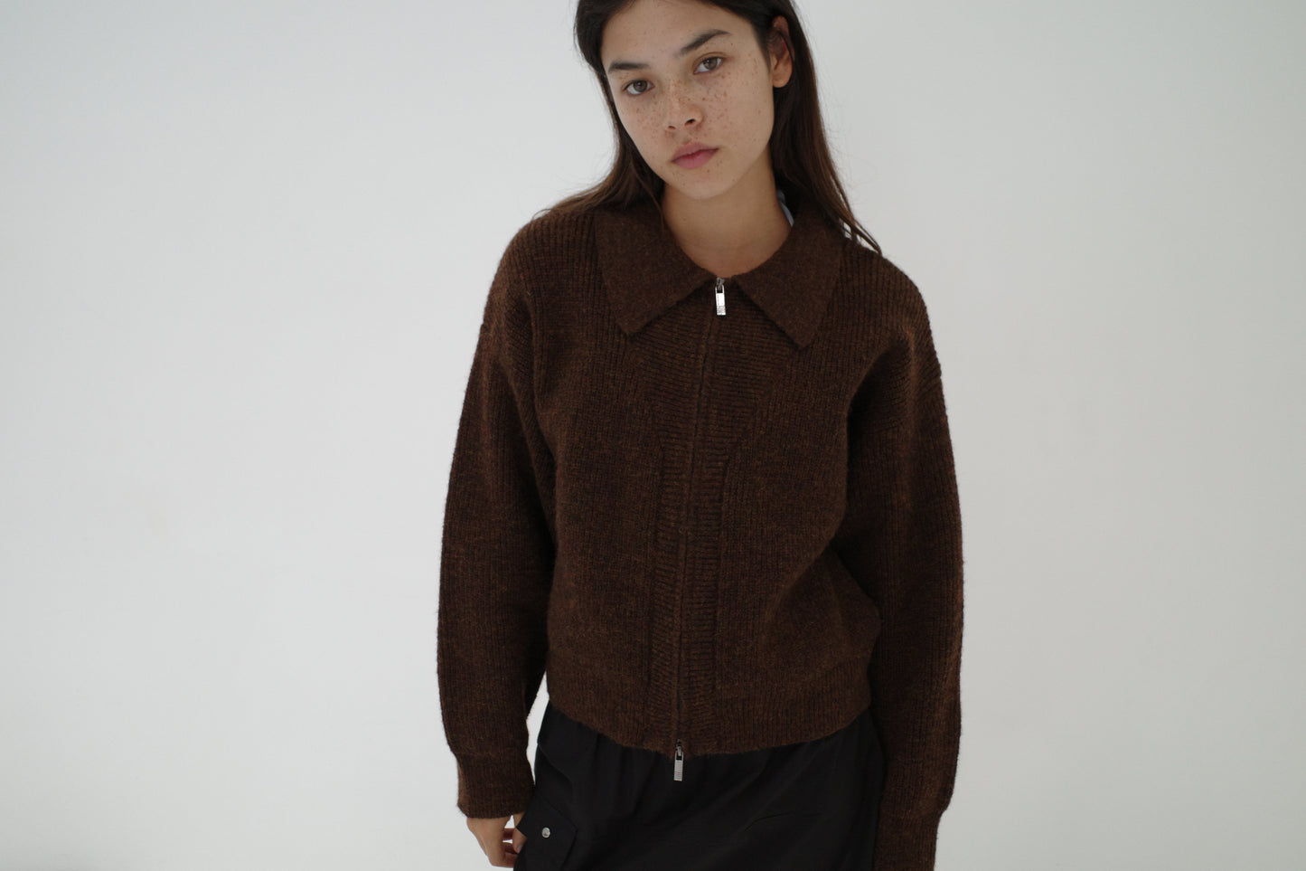 A young woman with long dark hair wears the Double Layer Cardigan - Brown from Deiji Studios and black pants, standing against a plain white background and looking slightly downward with a neutral expression.