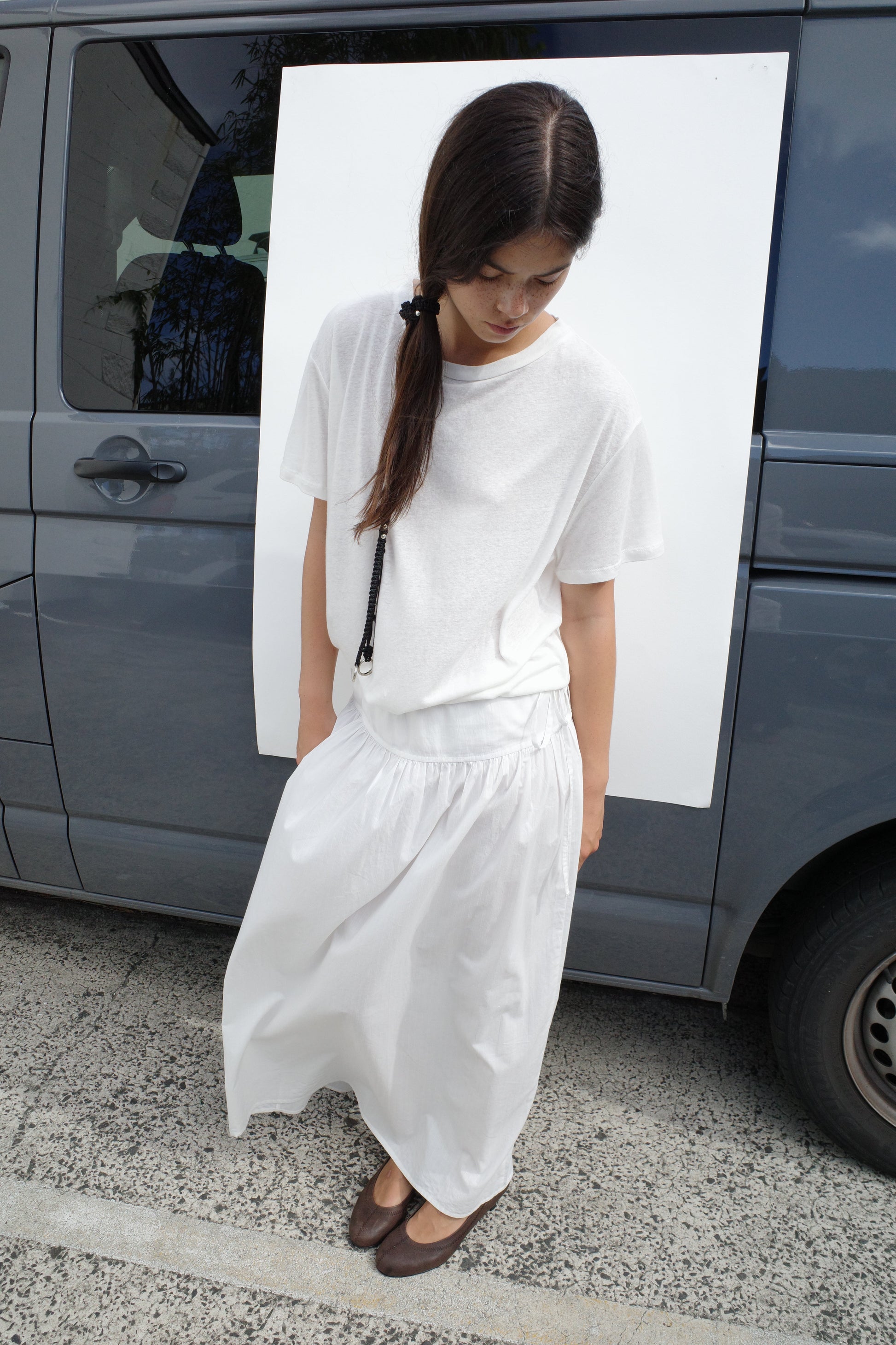 A woman with long dark hair in a side ponytail stands before a van, wearing a loose white T-shirt paired with the Maxi Skirt - White Texture by Deiji Studios and brown shoes, looking down with a neutral expression.