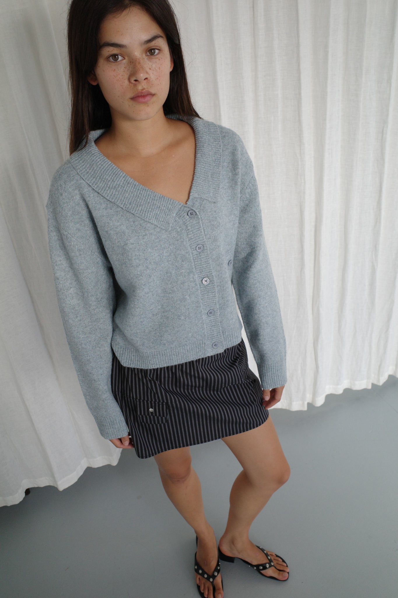 A woman stands indoors before white curtains, wearing Deiji Studios' Cross Button Cardigan in Ice Blue over a dark pinstriped skirt and black sandals. She has long brown hair, a knit layer for texture, and a neutral expression.
