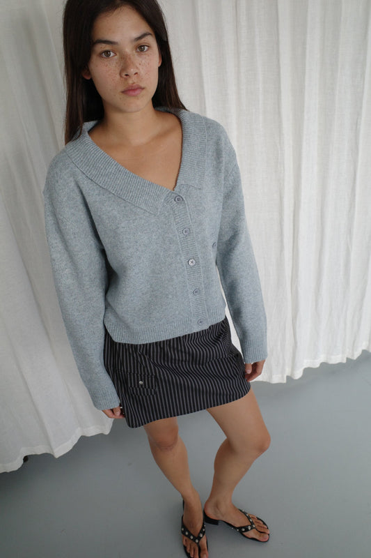 A woman stands indoors before white curtains, wearing Deiji Studios' Cross Button Cardigan in Ice Blue over a dark pinstriped skirt and black sandals. She has long brown hair, a knit layer for texture, and a neutral expression.
