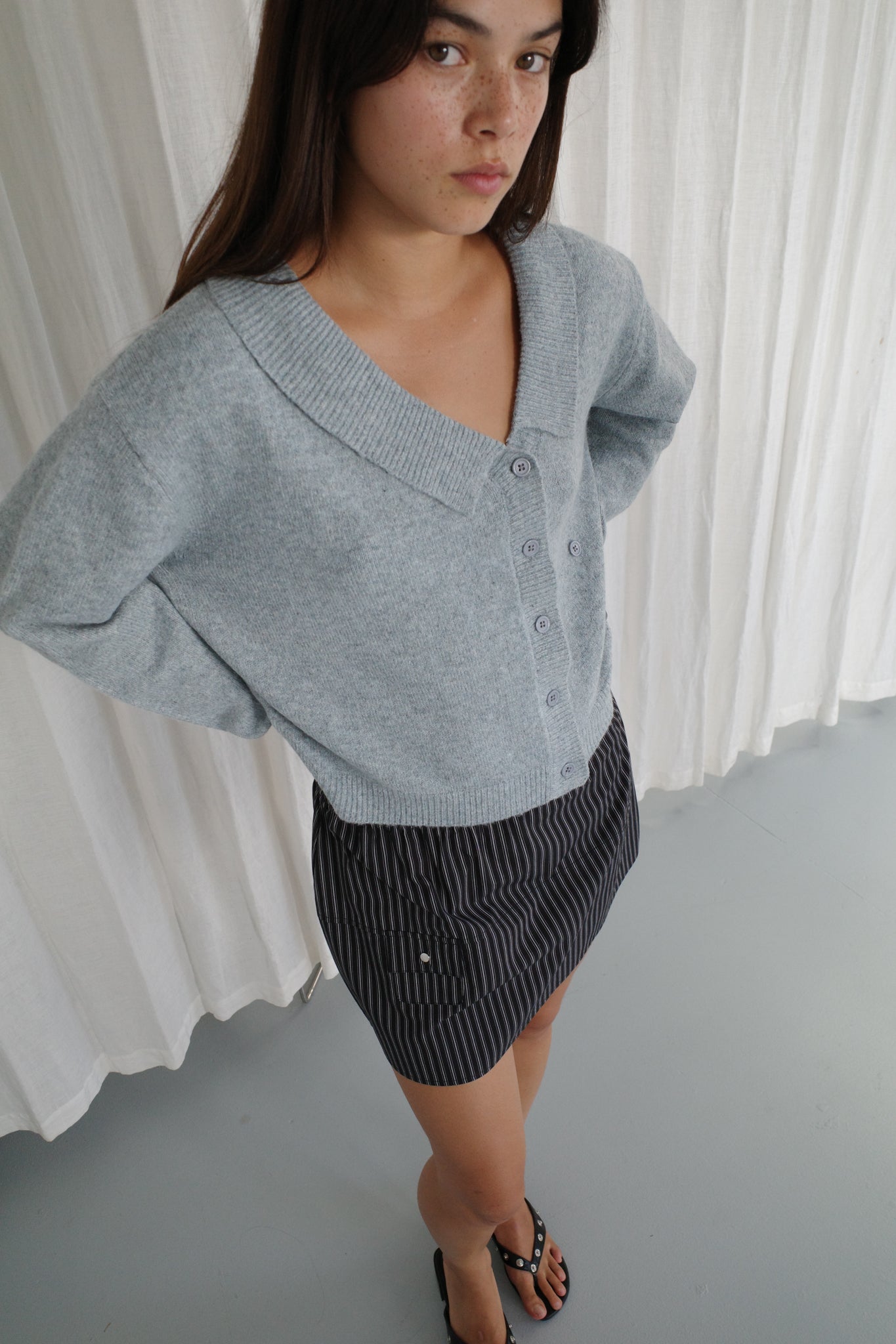 Young woman with long brown hair wears a Deiji Studios Cross Button Cardigan in Ice Blue as a knit layer, paired with a black and white pinstripe skirt and black sandals, standing indoors on a light gray floor with white curtains behind her.