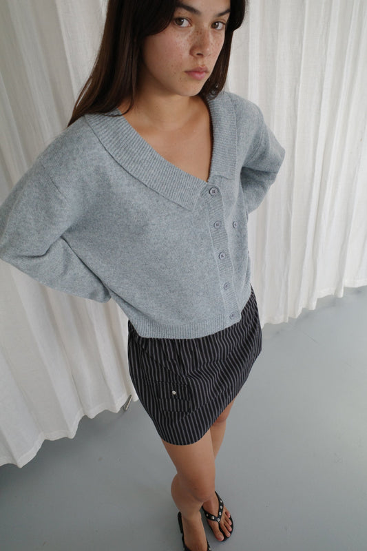Young woman with long brown hair wears a Deiji Studios Cross Button Cardigan in Ice Blue as a knit layer, paired with a black and white pinstripe skirt and black sandals, standing indoors on a light gray floor with white curtains behind her.