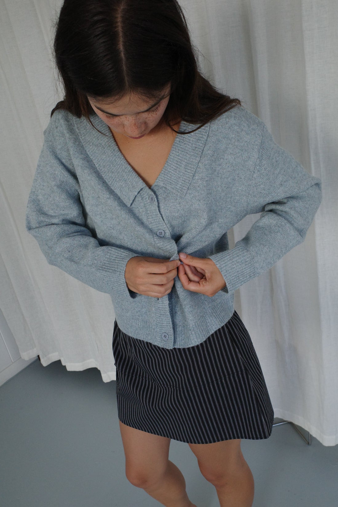 A person with long brown hair looks down while buttoning an Ice Blue Cross Button Cardigan by Deiji Studios. They wear a black skirt with white pinstripes and stand in front of white curtains.