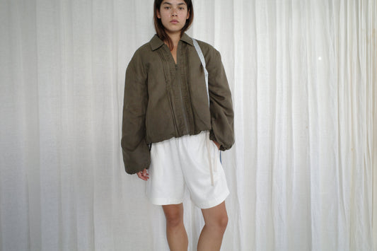 A person with straight, dark hair and a neutral expression stands before white curtains, wearing the Deiji Studios Pintuck Jacket - Khaki Stripe over a white outfit with knee-length shorts.