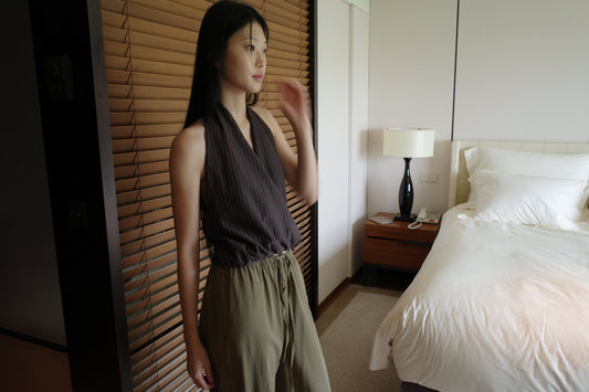 A woman with long dark hair stands beside wooden blinds in a bedroom wearing the Wrap Halter Top - Brown Grey Stripe by Deiji Studios, a sheer brown and grey striped halter top, with khaki trousers.