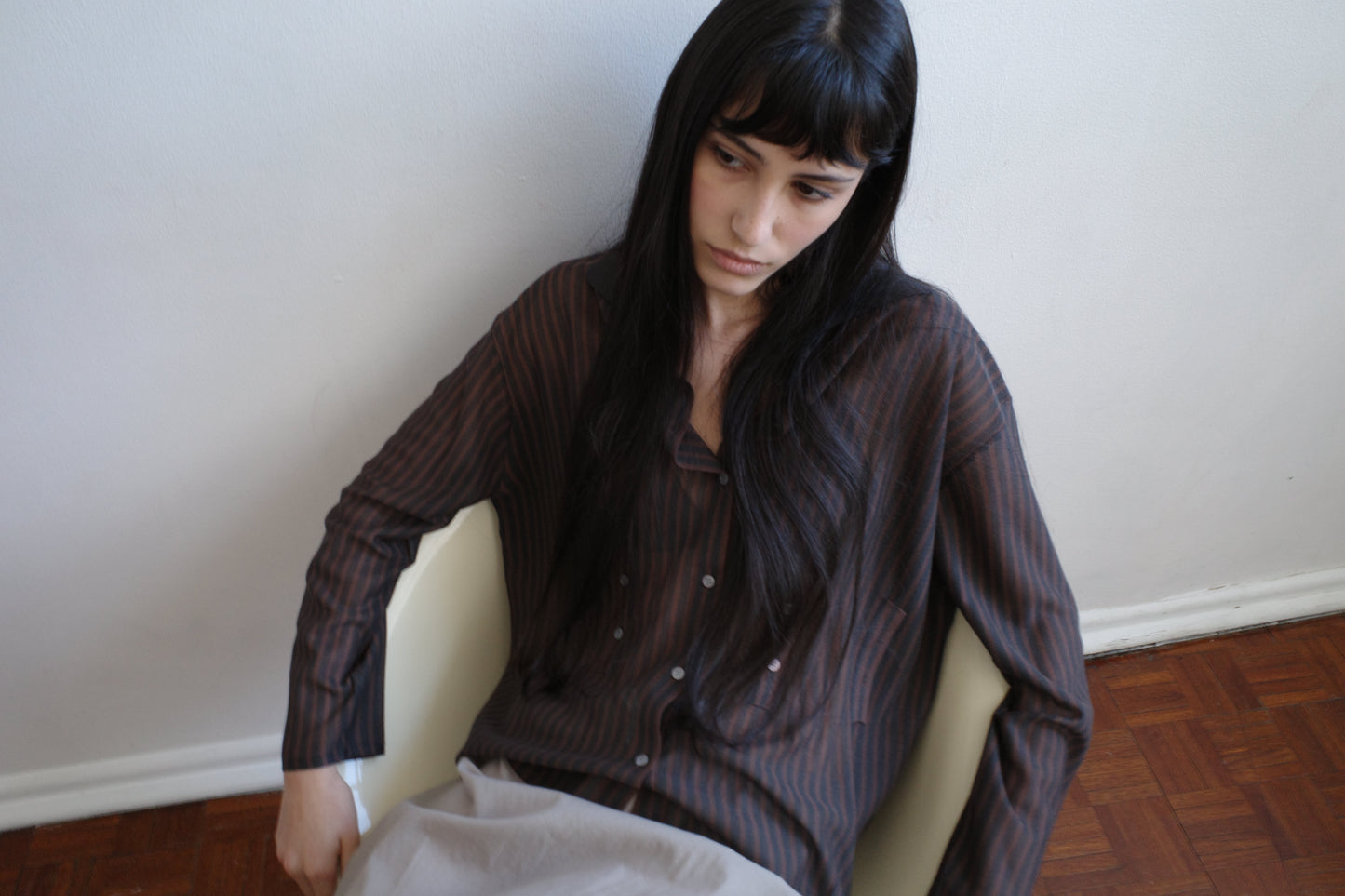A woman with dark hair and a fringe sits in a pale armchair looking down, wearing the Knit Shirt - Brown Grey Stripe by Deiji Studios, a sheer brown and charcoal striped knit shirt.