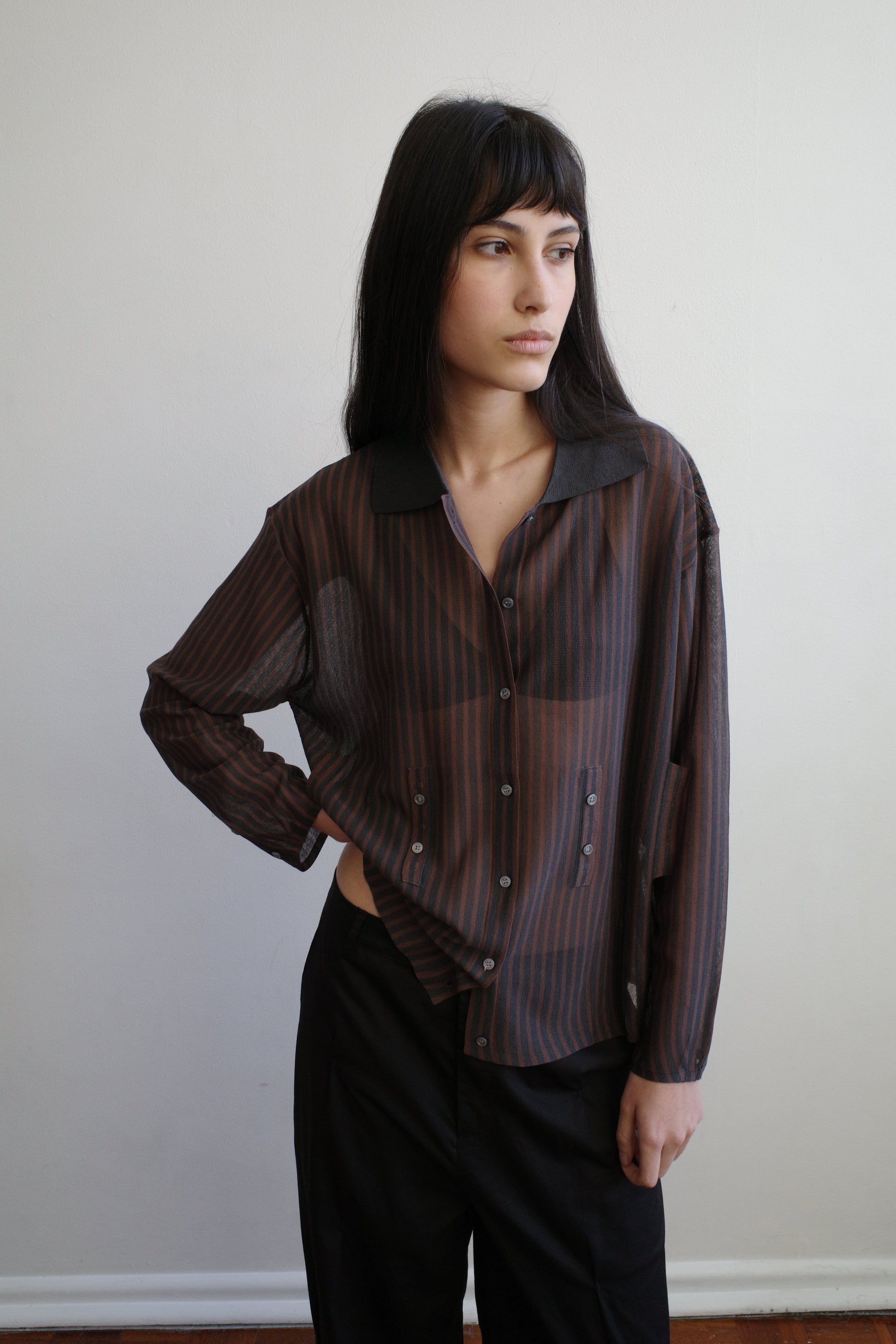 A woman with dark hair and a fringe stands against a pale wall wearing the Knit Shirt - Brown Grey Stripe by Deiji Studios, a sheer brown and charcoal striped knit shirt with a dark collar, over black trousers.