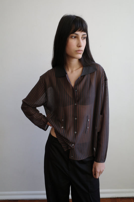A woman with dark hair and a fringe stands against a pale wall wearing the Knit Shirt - Brown Grey Stripe by Deiji Studios, a sheer brown and charcoal striped knit shirt with a dark collar, over black trousers.