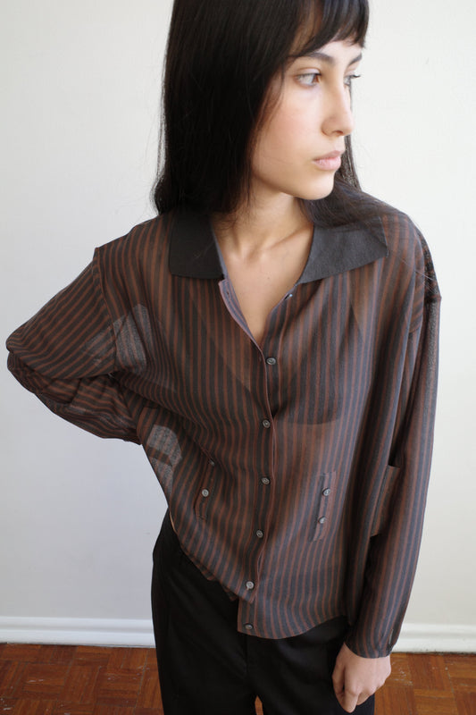 A woman with dark hair and a fringe stands against a pale wall wearing the Knit Shirt - Brown Grey Stripe by Deiji Studios, a sheer brown and charcoal striped knit shirt with a dark collar, over black trousers.