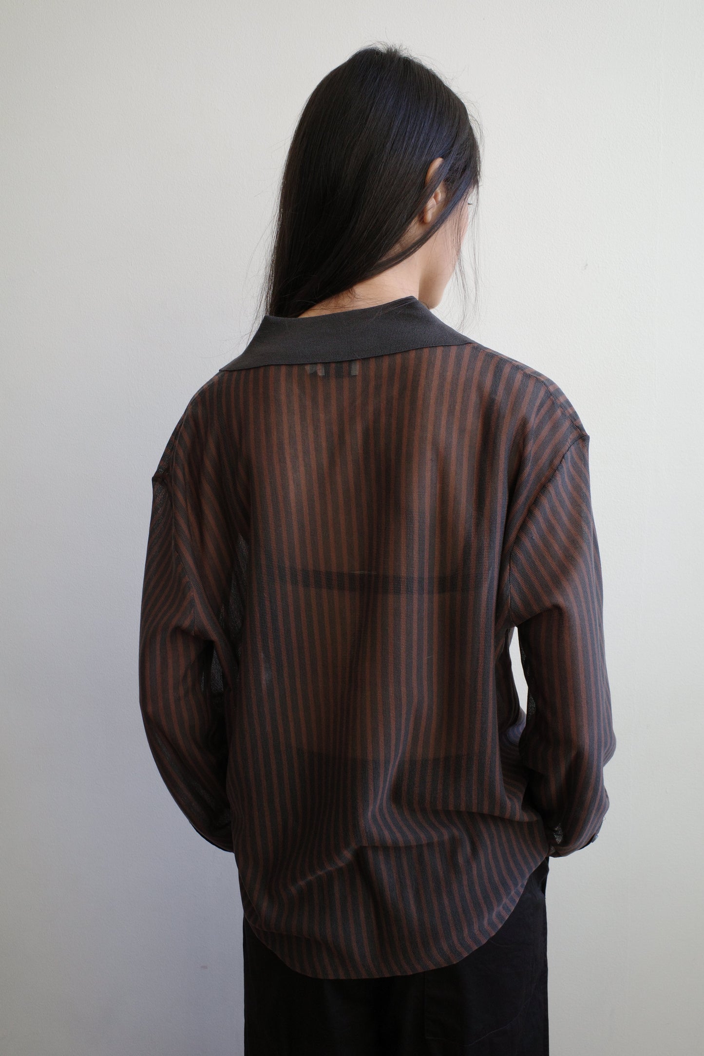 A woman with long dark hair stands with her back to the camera against a pale wall wearing the Knit Shirt - Brown Grey Stripe by Deiji Studios, a sheer brown and charcoal striped knit shirt with a dark collar.