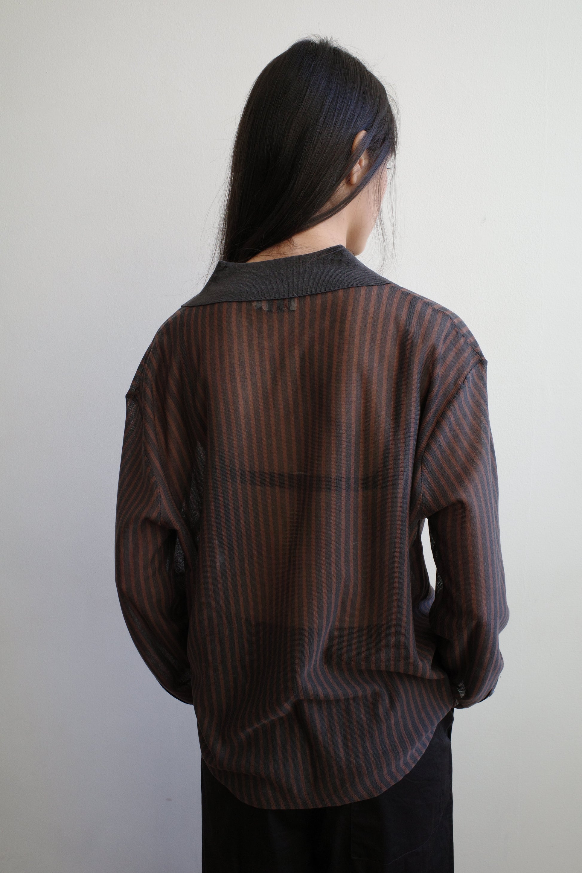 A woman with long dark hair stands with her back to the camera against a pale wall wearing the Knit Shirt - Brown Grey Stripe by Deiji Studios, a sheer brown and charcoal striped knit shirt with a dark collar.
