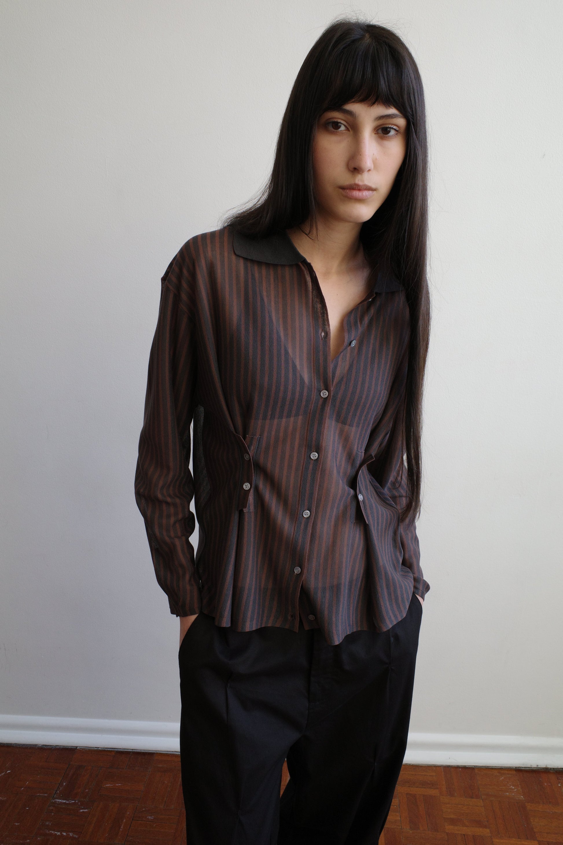 A woman with long dark hair stands against a pale wall wearing the Knit Shirt - Brown Grey Stripe by Deiji Studios, a sheer brown and charcoal striped knit shirt with a dark collar, over black trousers.
