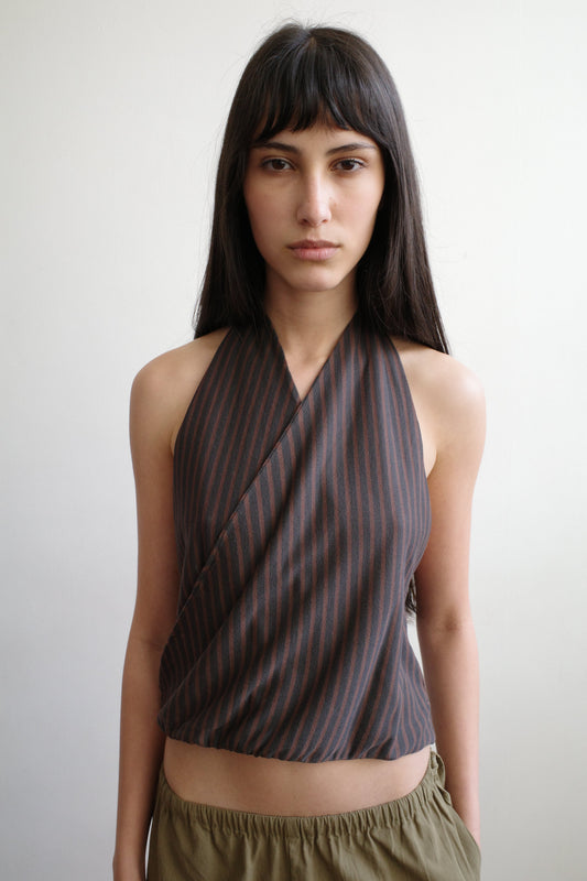 A cropped front view of a woman with a fringe wearing the Wrap Halter Top - Brown Grey Stripe by Deiji Studios, a sheer brown and grey striped halter top with a wrap surplice front, with khaki trousers.