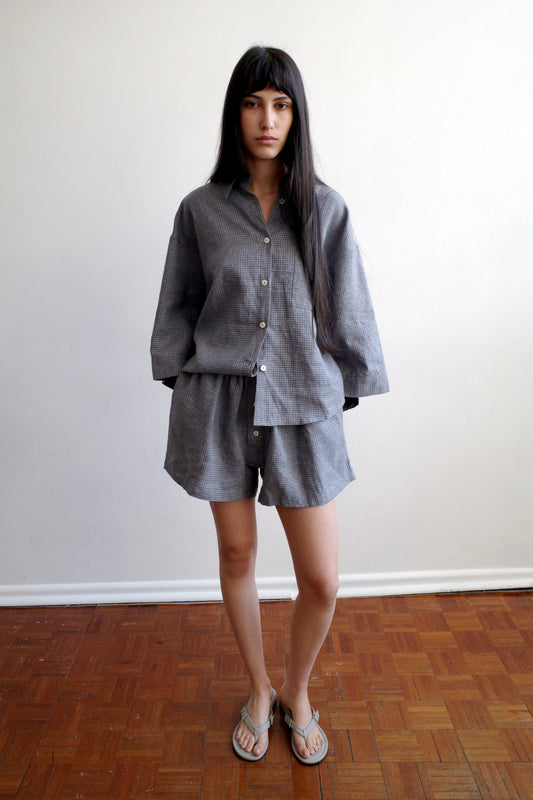 A woman stands indoors on a wooden floor, wearing the 03 Set - Indigo Check from Deiji Studios—featuring a loose linen shirt and matching boxer style shorts—and flip-flops, set against a plain white wall.