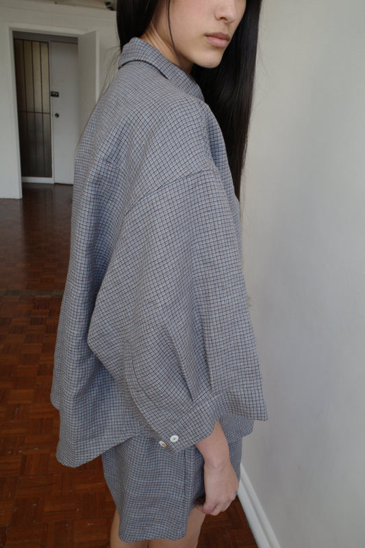 A woman with long dark hair and a fringe stands facing the camera in a room, wearing the 03 Set - Indigo Check by Deiji Studios, an oversized boxy check linen shirt with matching boxer-style shorts.