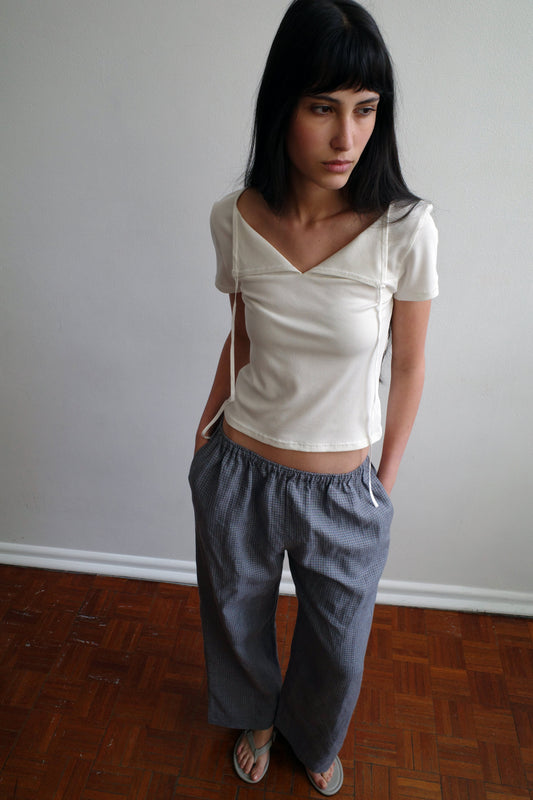 A woman with dark hair and a fringe stands against a pale wall wearing the Tie Back Tee - White by Deiji Studios, a fitted cream tee with a sweetheart neckline, with grey check trousers and flip-flops.