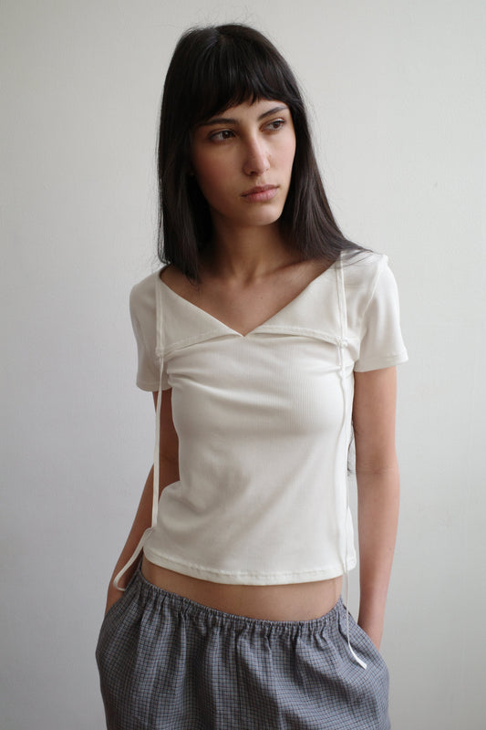 A woman with dark hair and a fringe stands against a pale wall wearing the Tie Back Tee - White by Deiji Studios, a fitted cream tee with a sweetheart neckline and back ties, with grey check trousers.