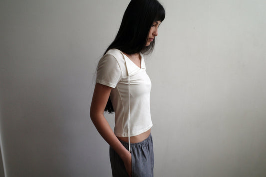 A woman with dark hair and a fringe stands in profile against a pale wall wearing the Tie Back Tee - White by Deiji Studios, a fitted cream tee with a sweetheart neckline and back tie, with grey check trousers.