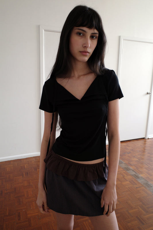A woman with dark hair and a fringe stands in a room wearing the Tie Back Tee - Black by Deiji Studios, a fitted black tee with a sweetheart neckline and back ties, with a brown flounce mini skirt.