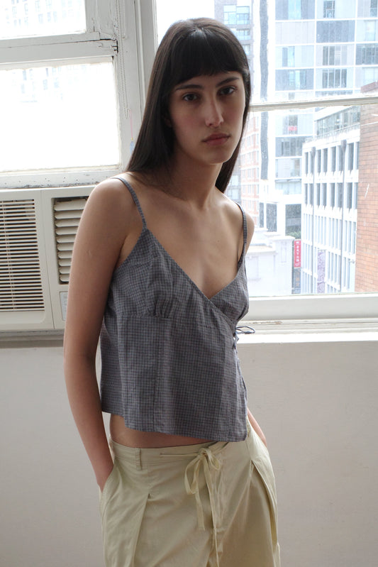 A woman with dark hair and a fringe stands by a window wearing the Wrapping Top - Indigo Check by Deiji Studios, an indigo micro-check linen cami with a wrap front, and cream trousers.