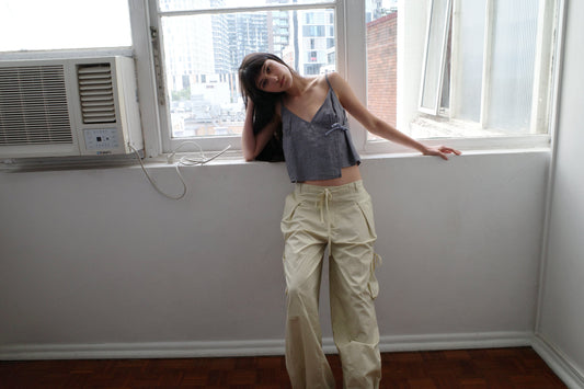 A woman with dark hair and a fringe leans against a window sill wearing the Wrapping Top - Indigo Check by Deiji Studios, an indigo micro-check linen cami, with cream trousers.
