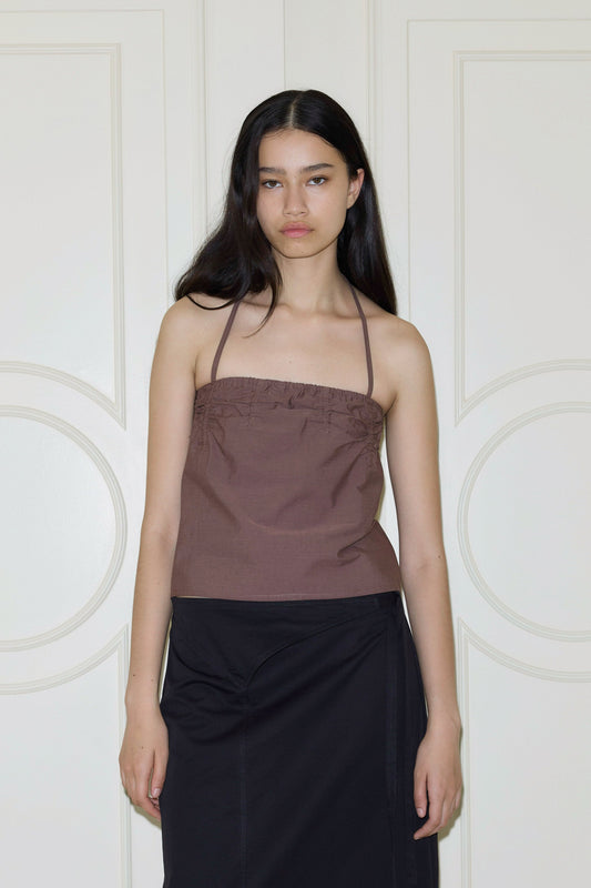 A woman with long dark hair wears a brown Deiji Studios Tube Top and a black skirt, standing before white paneled doors and looking at the camera with a neutral expression.