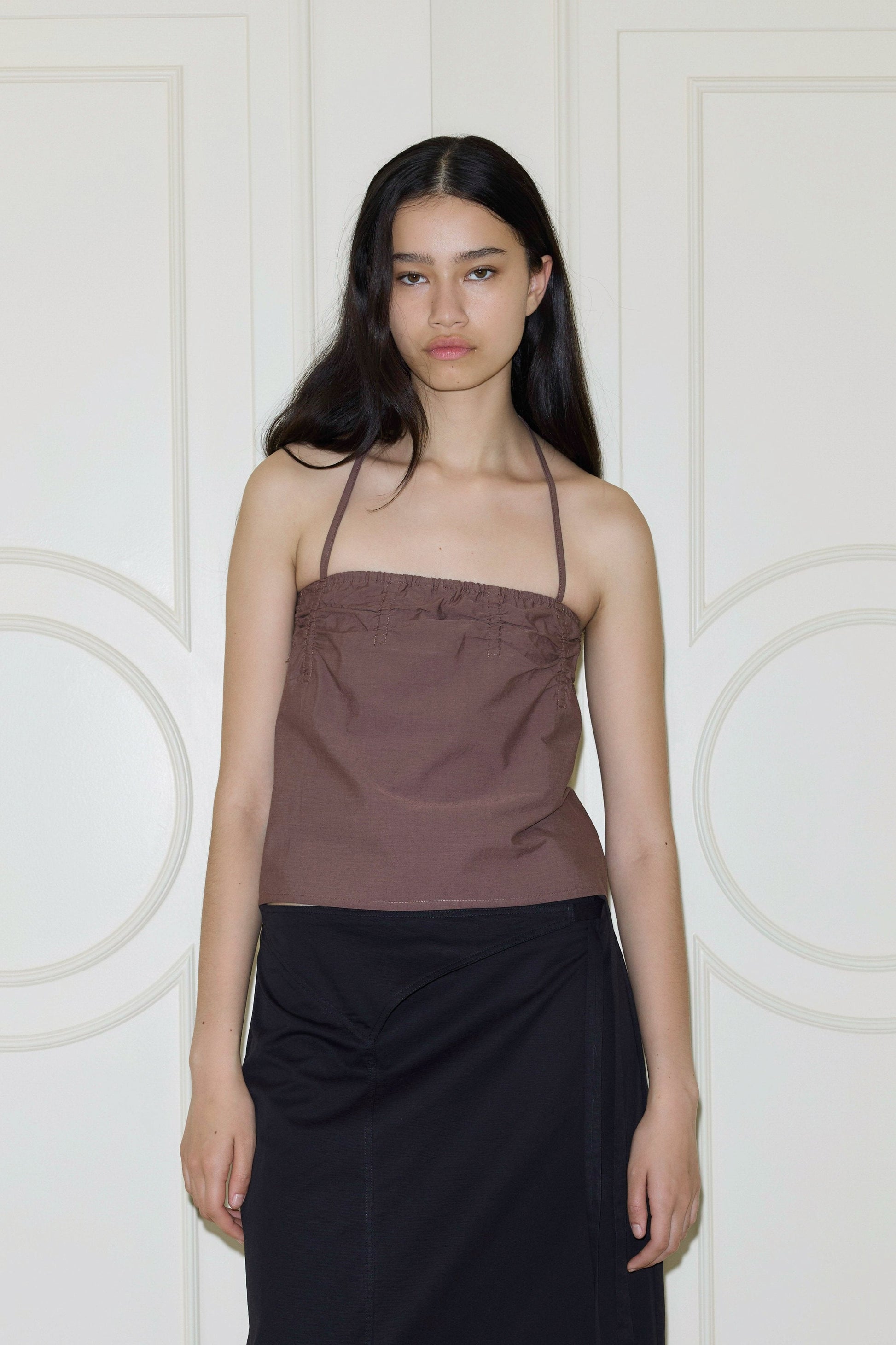 A woman with long dark hair wears a brown Deiji Studios Tube Top and a black skirt, standing before white paneled doors and looking at the camera with a neutral expression.