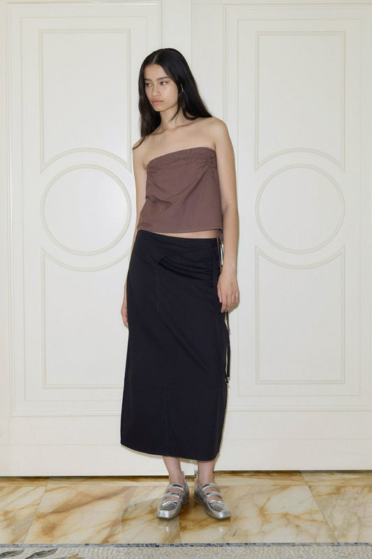 A woman with long dark hair wears a brown strapless top, silver sandals, and the Crossover skirt in black. She stands indoors on a marble floor before white paneled doors, gazing slightly to the side.