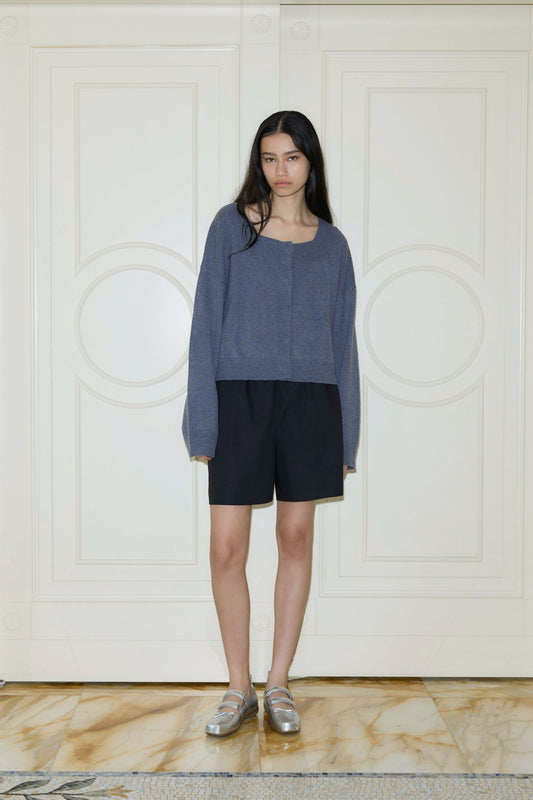 A young woman with long dark hair stands indoors by white paneled doors, wearing a loose gray sweater, silver loafers, and the Button Short on a marble floor.