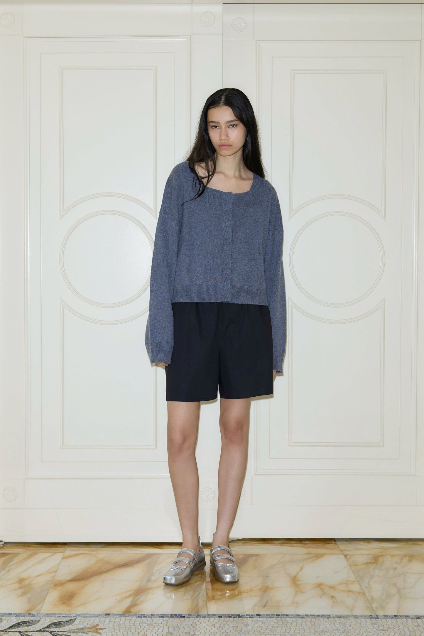 A young woman with long dark hair stands indoors before cream paneled doors, wearing the Button Short with a loose gray sweater and silver loafers, looking at the camera with a neutral expression.