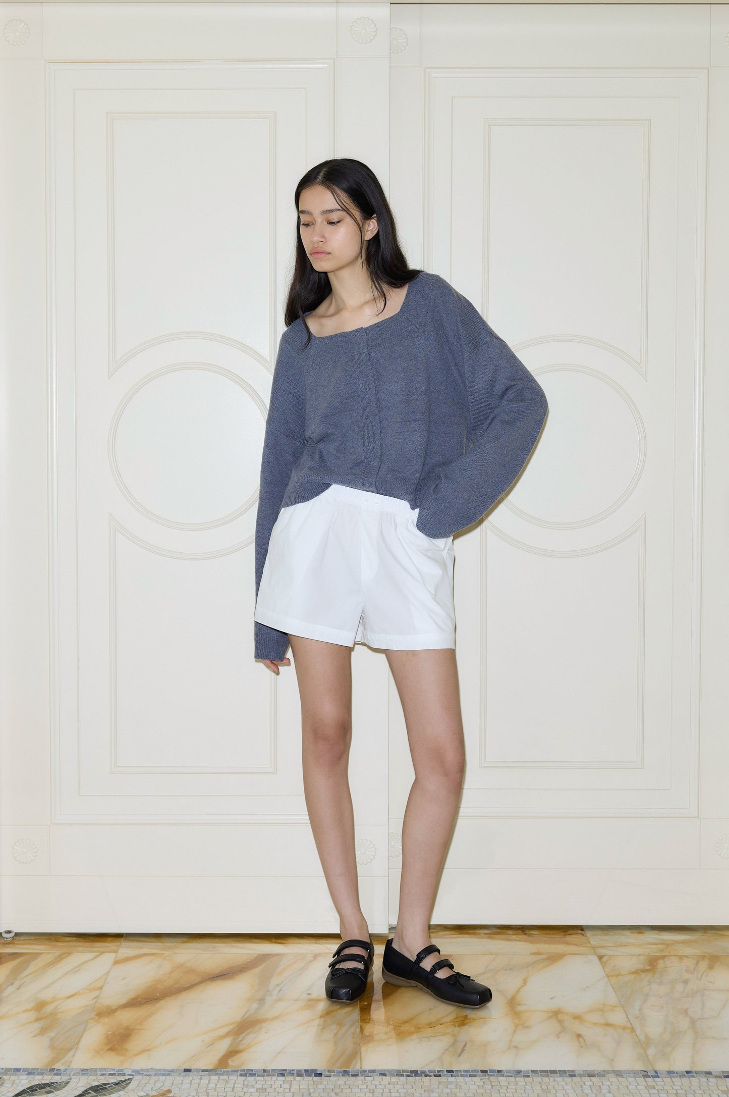 A young woman with long dark hair stands indoors against a cream-paneled wall, wearing Deiji Studios Organic Cotton Boxer shorts and a loose gray sweater, paired with black shoes for a relaxed look.