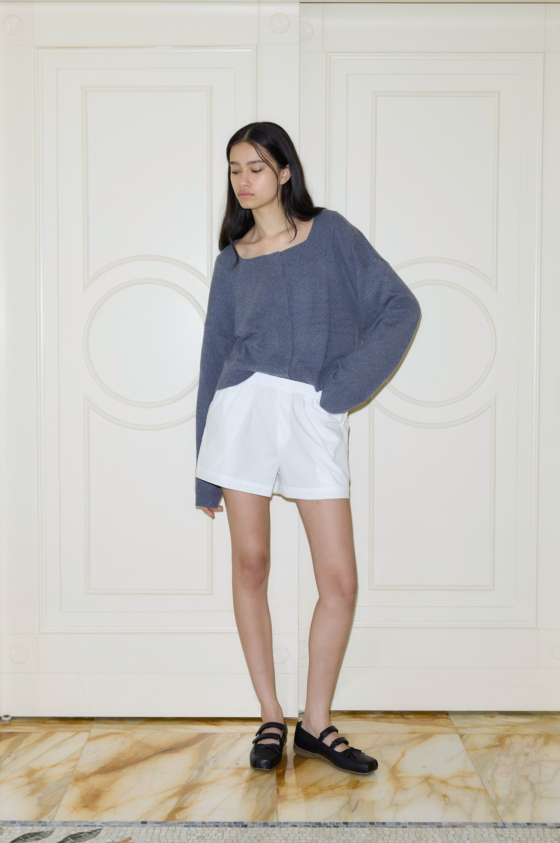 A young woman with long dark hair stands indoors against a cream-paneled wall, wearing Deiji Studios Organic Cotton Boxer shorts and a loose gray sweater, paired with black shoes for a relaxed look.