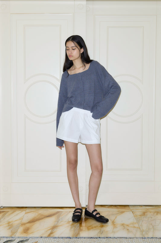A young woman with long dark hair stands indoors against a cream-paneled wall, wearing Deiji Studios Organic Cotton Boxer shorts and a loose gray sweater, paired with black shoes for a relaxed look.