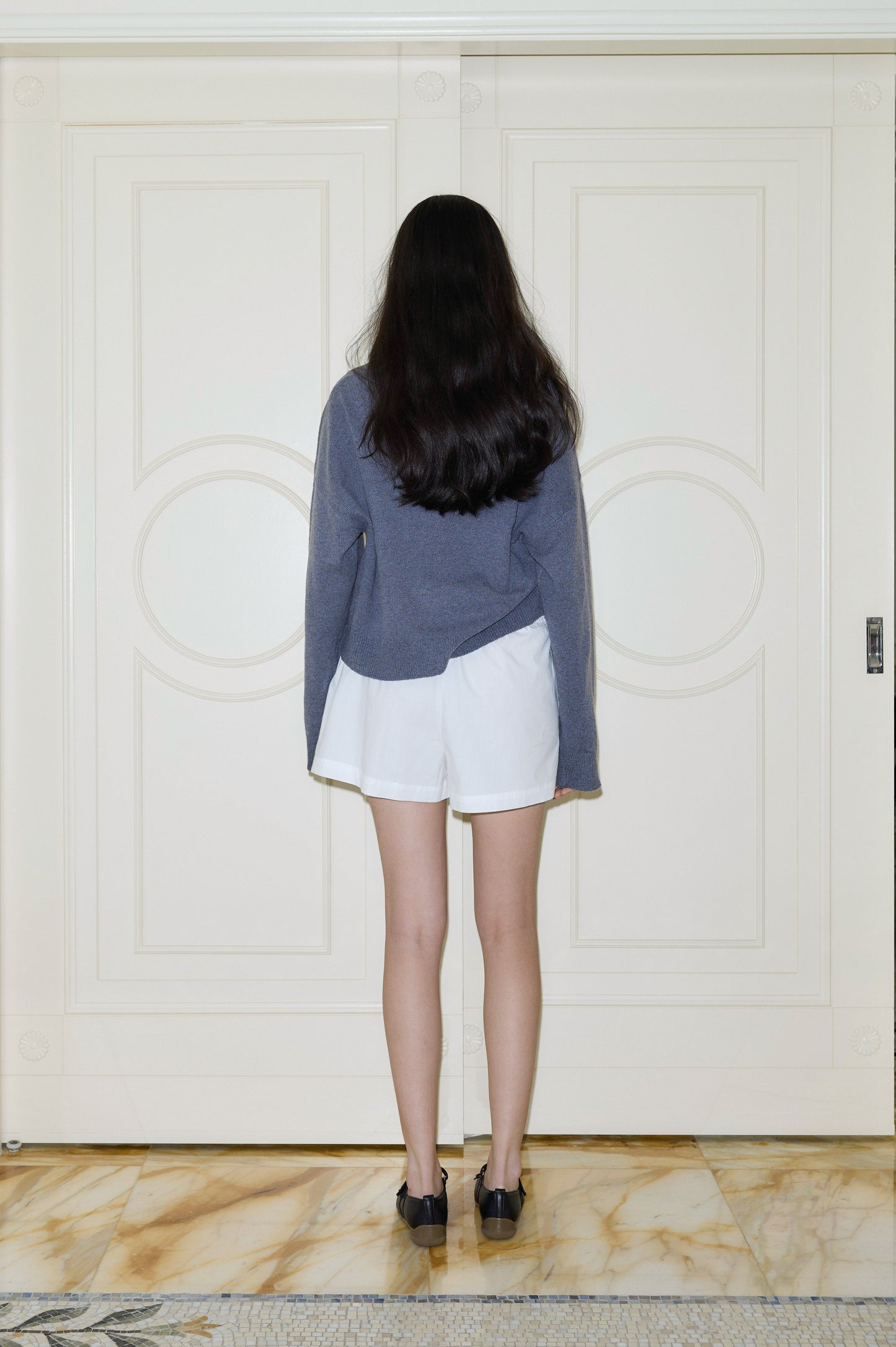 A person with long, dark hair, dressed in a blue long-sleeve shirt, black shoes, and the Organic Cotton Boxer - White, stands on a marble floor facing a closed white double door. The individual is viewed from behind.