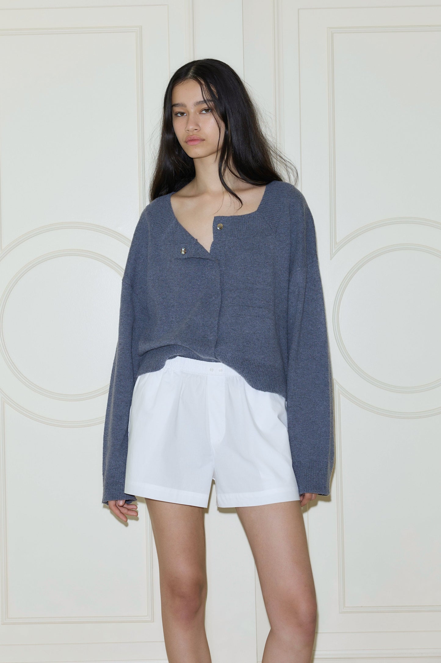 A woman with long dark hair stands against a cream wall, wearing the Organic Cotton Boxer - White and a loose blue-gray buttoned sweater, looking slightly to the side with a neutral expression.