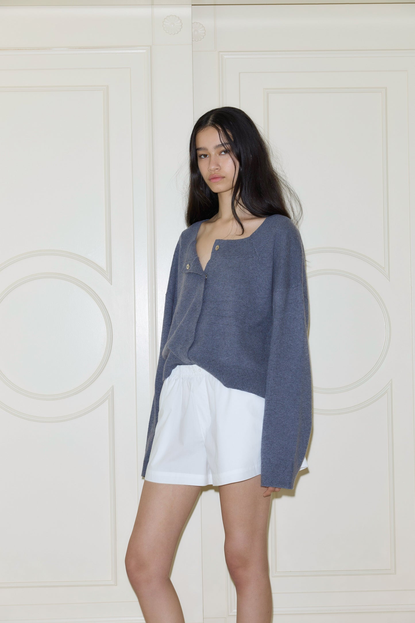 A woman with long dark hair stands indoors against a cream paneled wall, wearing a loose blue sweater and Deiji Studios Organic Cotton Boxer in White. She faces the camera with a neutral expression.