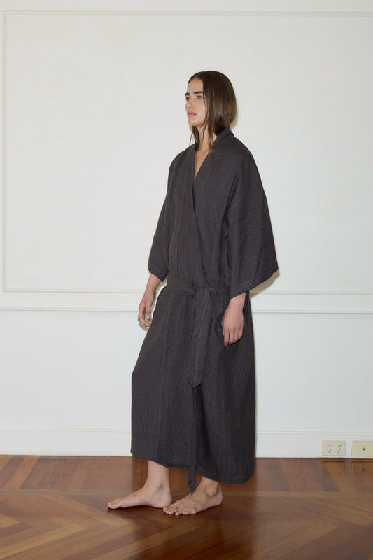 A person with long brown hair stands barefoot on a wooden floor, wearing the 02 Robe—a dark, loose-fitting garment with wide sleeves and a waist tie—against a plain white wall.